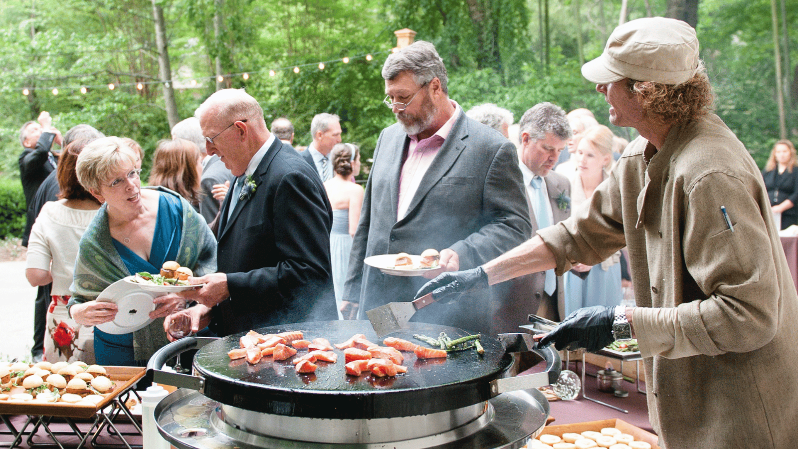 Chef in tan hat and chef's coat is at the Evo ventless tabletop action station. On the grill area pieces of fish. In front of table are three guests, two men and one woman, looking at the chef. Guests are dressed in fancy clothes. 