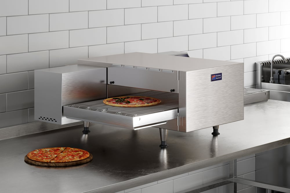 Silver Middleby Marshall conveyor oven sits on stainless steel countertop. One pizza on counter on side of oven when while another pizza is on belt. 