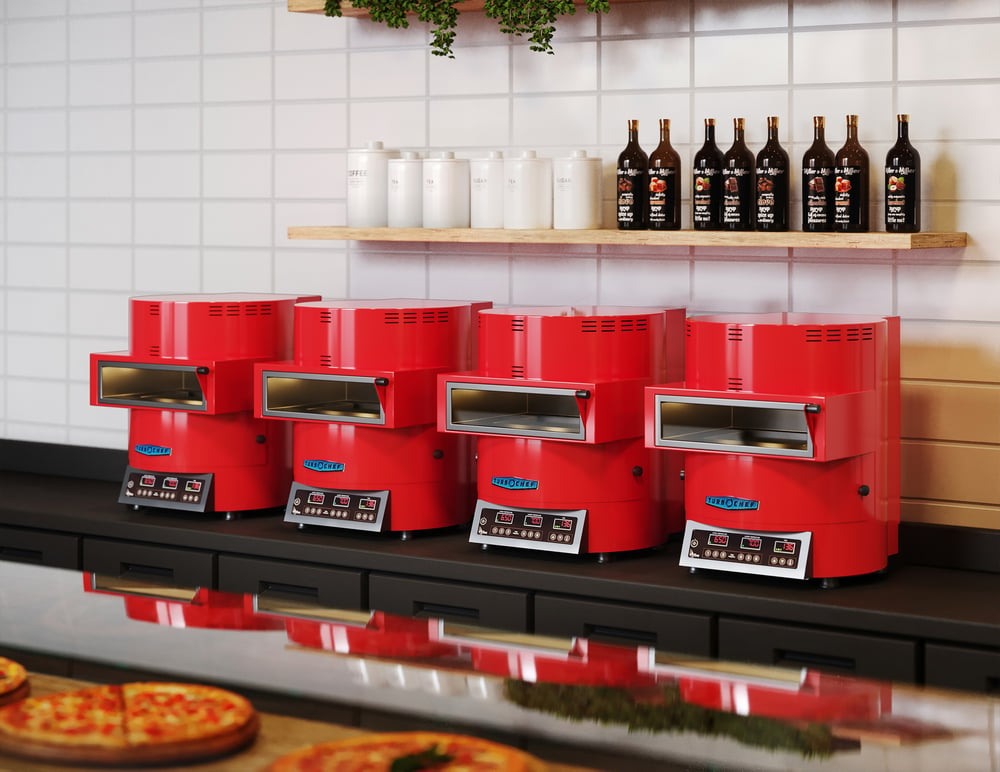 Four red TurboChef ventless Fire ovens sit on counter. Above on shelves are canisters and bottle. On counter in front of ovens are personal pizzas.