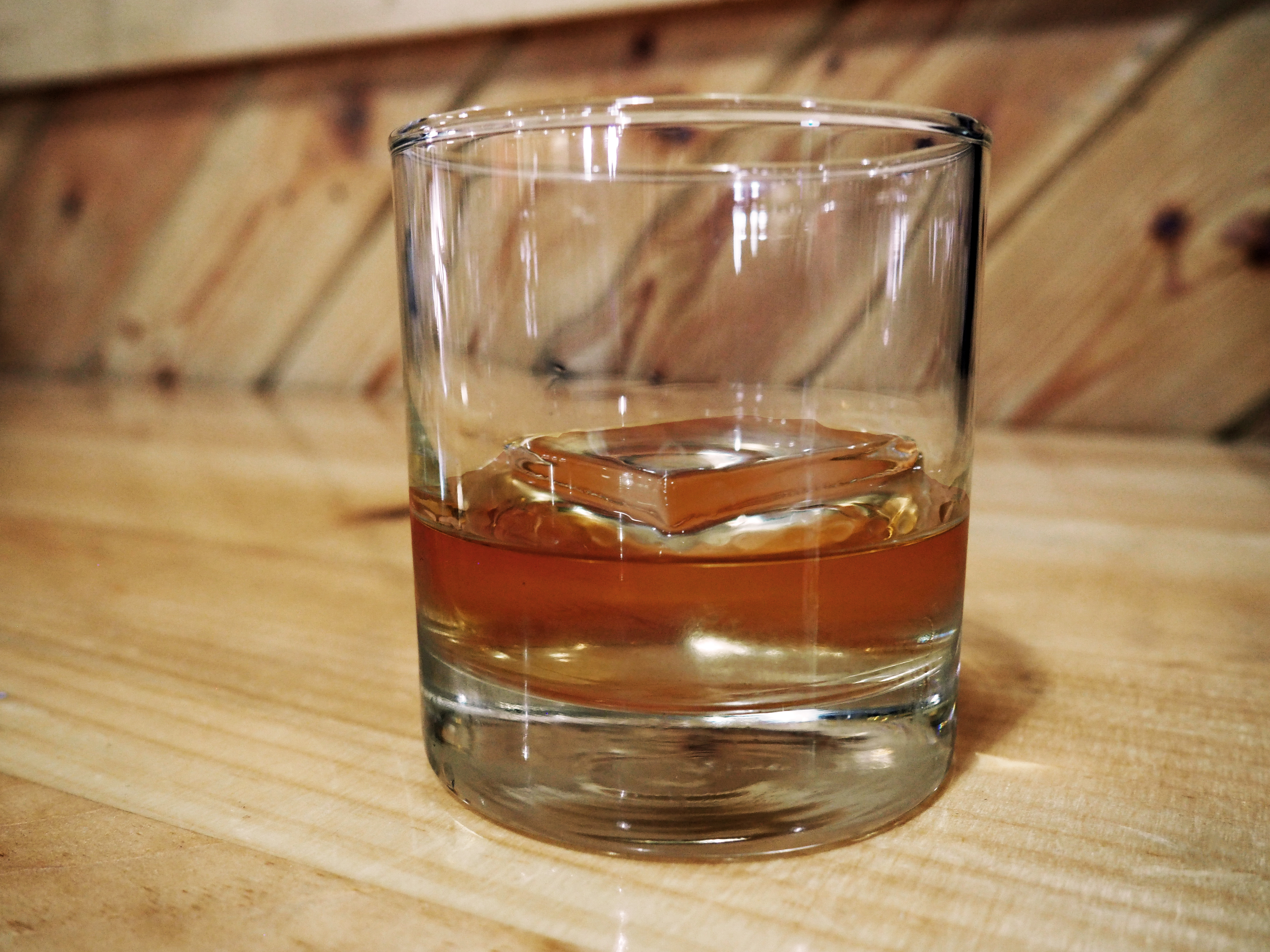 Cocktail glass with brown liquor and Viking clear ice cube inside. Glass is sitting on wood countertop with wood backsplash.